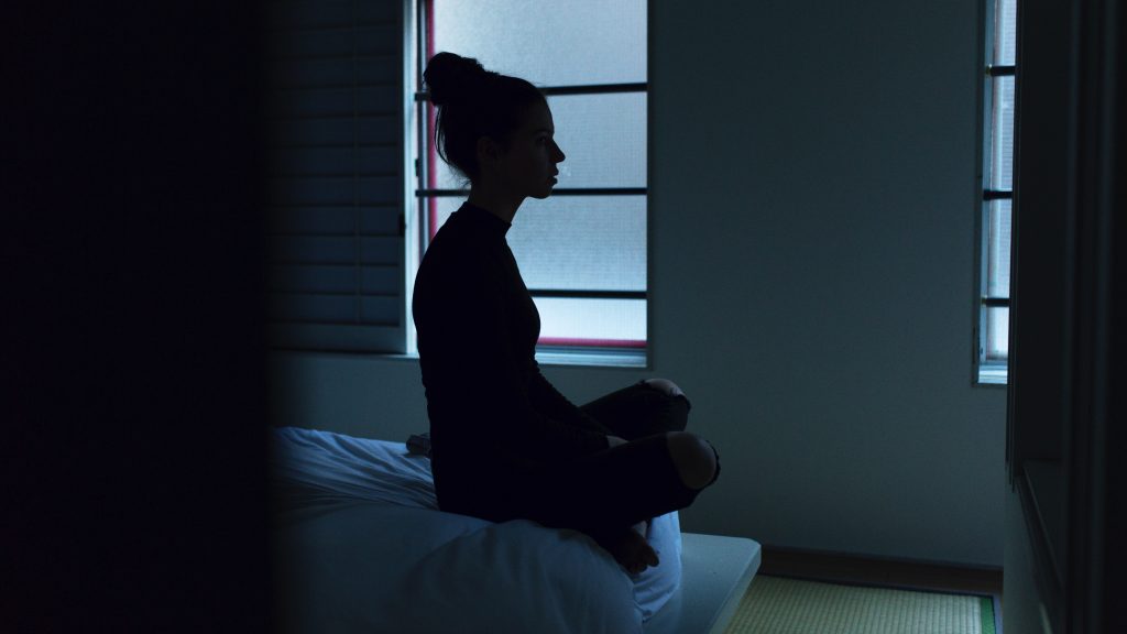 Silhouette of a woman meditating on bed in a low-light room.
