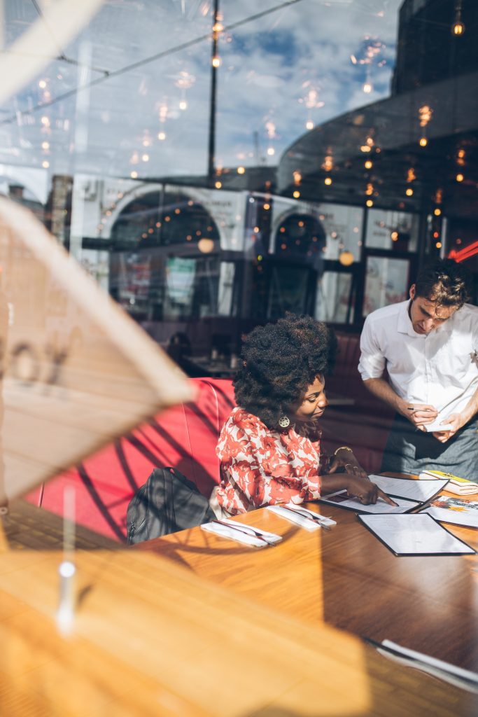 A woman points at a menu item as a waiter leans over to answer her question or take her order. 